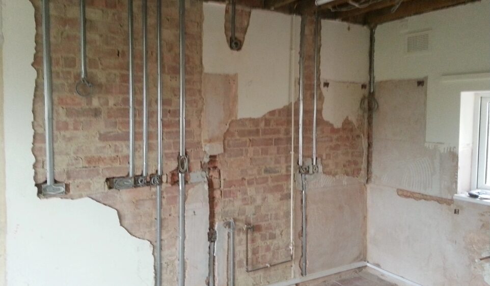 Exposed brick wall with newly installed metal electrical conduit and back boxes during a domestic rewiring project in a partially stripped residential room.
