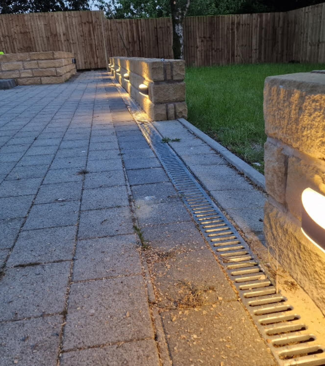 Evening view of a paved patio with integrated drainage channel and low stone wall, illuminated by built-in LED wall lights, next to a grassy lawn and wooden fence.