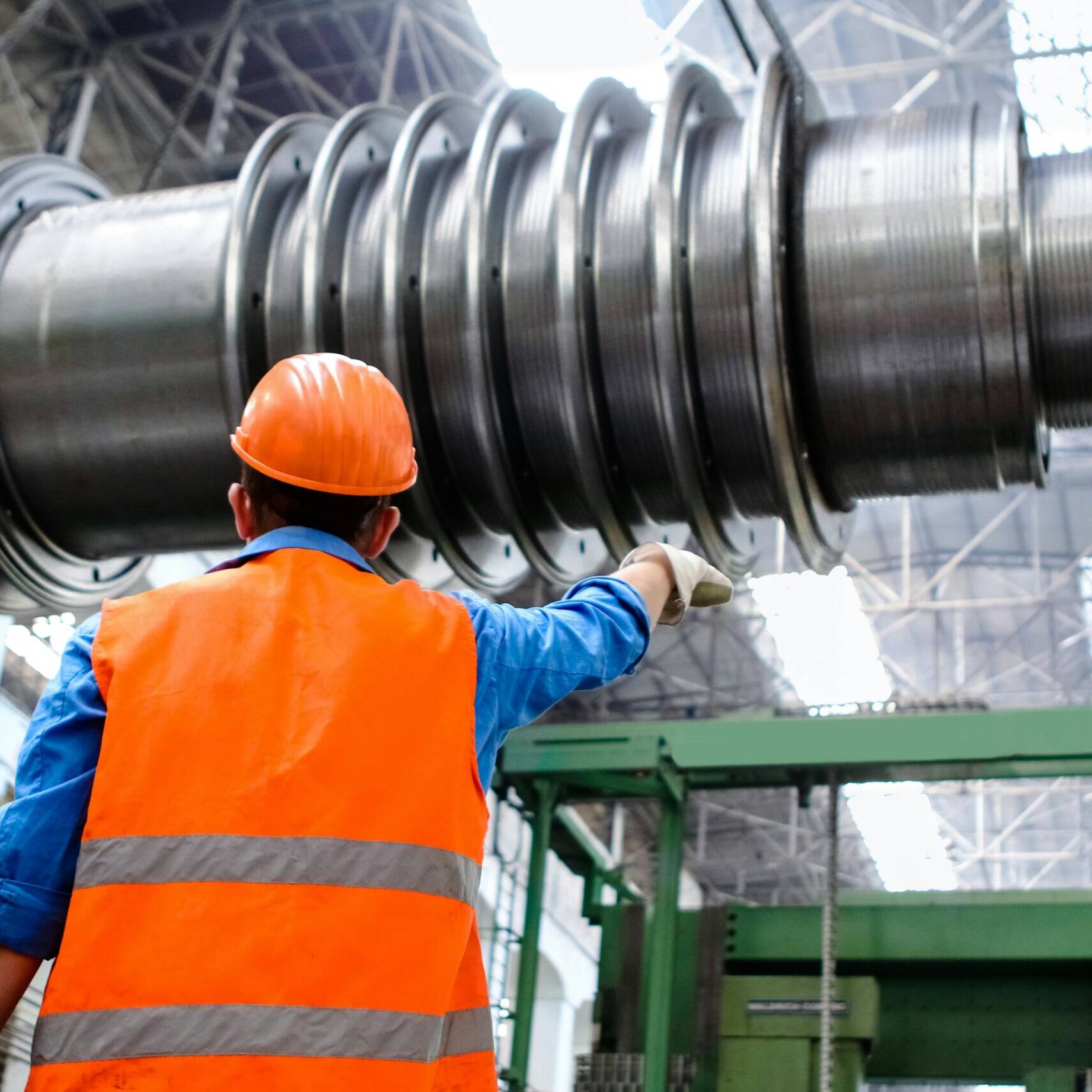 A factory worker in a orange hi-vis and hard hat, pointing at a piece of equipment.