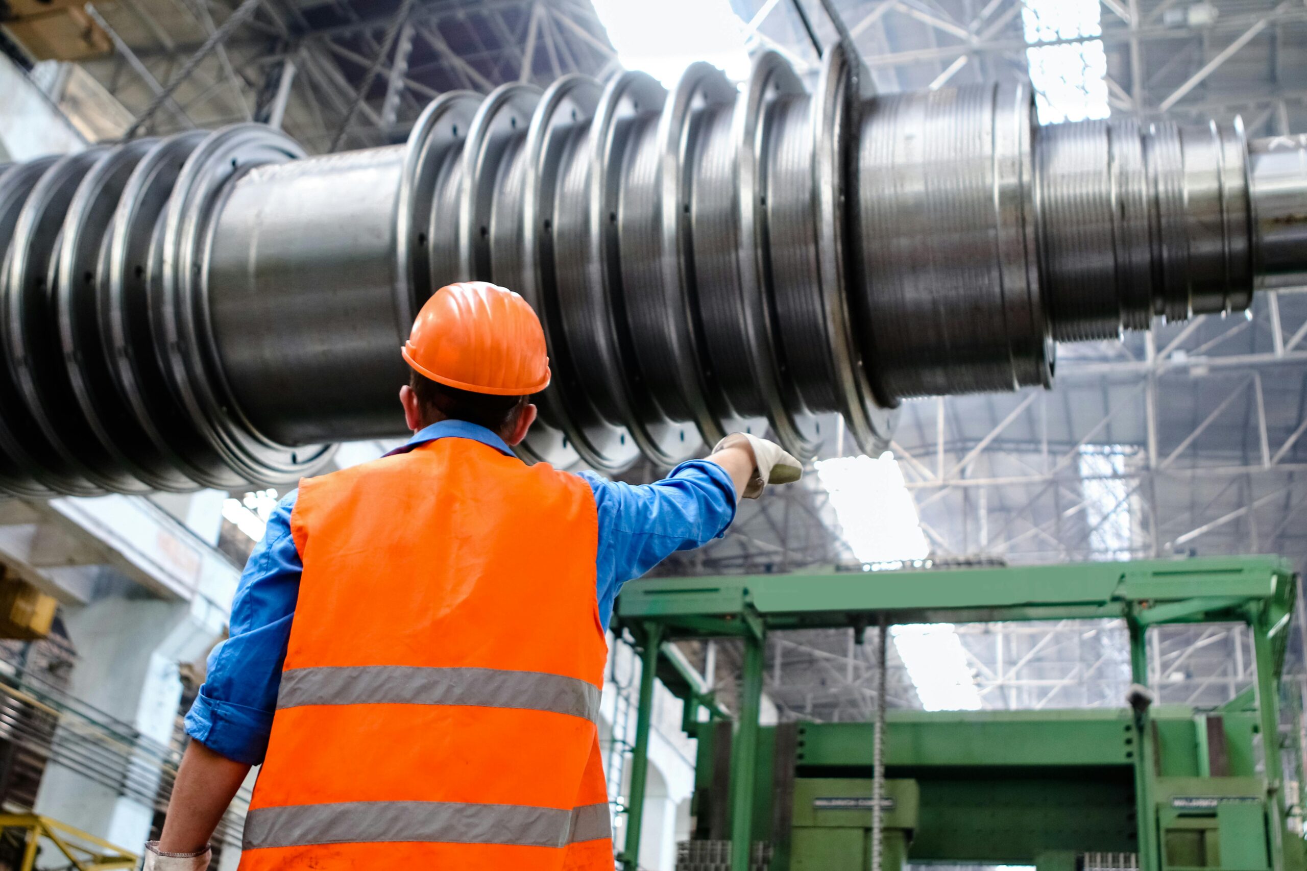 A factory worker in a orange hi-vis and hard hat, pointing at a piece of equipment.