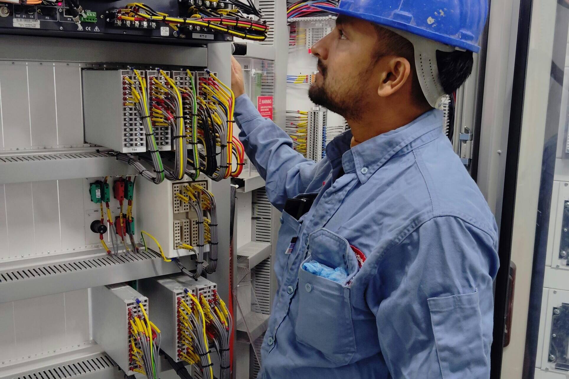 A man in a blue top and blue hard hat looking into a power box