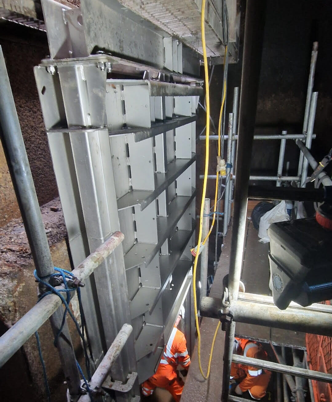 Engineers in high-visibility clothing working in a confined underground space, installing or inspecting a large metal cable tray or trunking system, surrounded by scaffolding and temporary lighting.