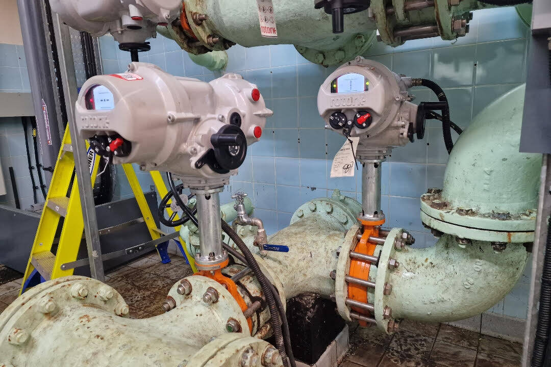 Industrial piping system with mounted electric actuators from Rotork in a tiled utility room. The setup includes large green pipes with orange flanges, control wiring, and a yellow ladder in the background, indicating ongoing maintenance or inspection work.