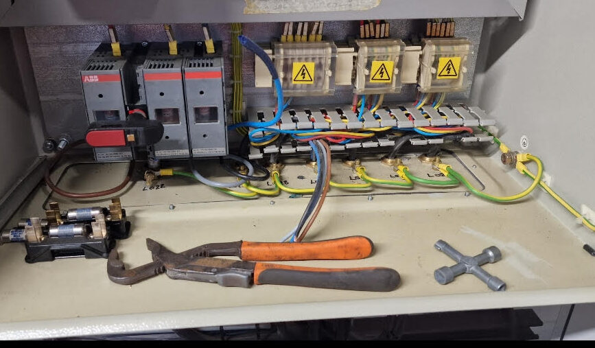 Interior of an electrical control panel showing circuit breaker, contractors, wiring and warning labels. Hand tools including pliers and a multi-purpose utility key are placed inside the open panel enclosure.